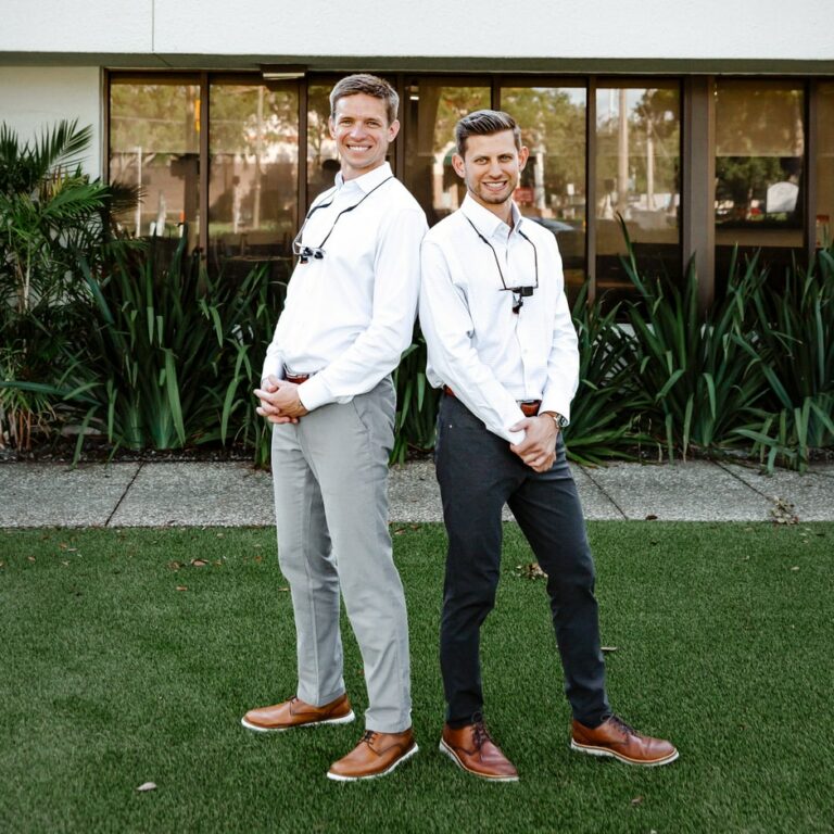 Dr. Rick Escobar and Dr. Clayton McEntire are joyfully smiling infront of Amley McEntire Orthodontics dentistry building