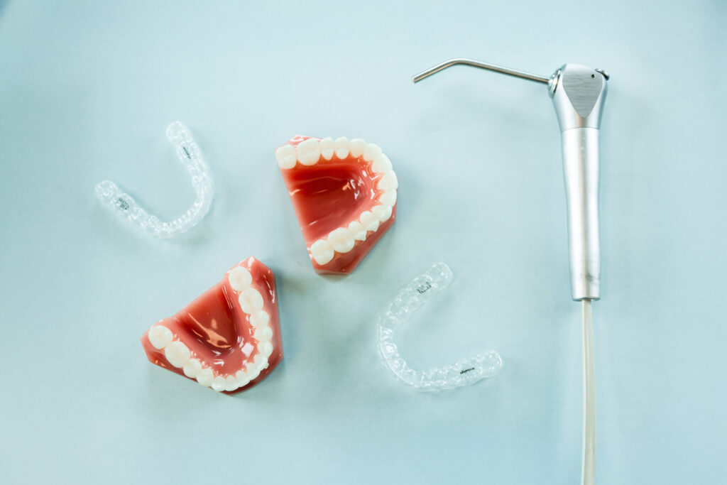 Two clear aligners, two dental models, and a dental tool on a flat table against a blue background.